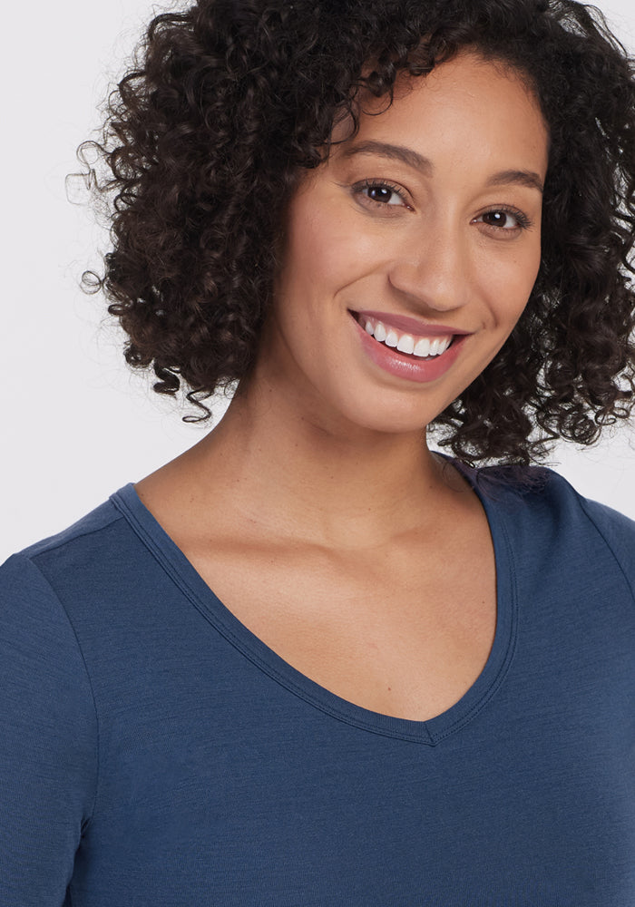 A woman with curly hair smiles at the camera, wearing the Woolx Layla V Neck Top in Starry Night. With minimal makeup and a natural look, she stands against a plain, light-colored background.