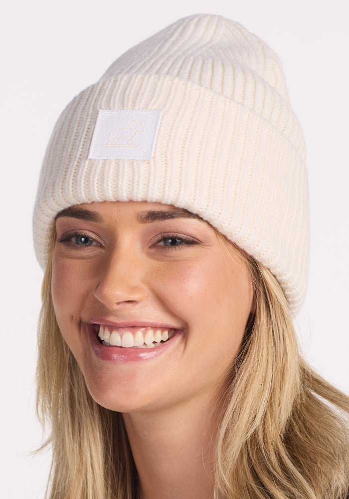 A young woman with long blonde hair smiles while wearing the Woolx Ria Chunky Beanie in Buttercream, a cozy winter accessory featuring a white front patch. The background is plain and light-colored.