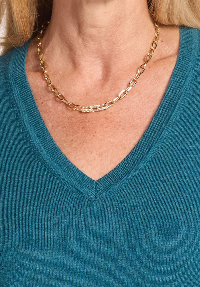 A person wears the Woolx Francesca Sweater in Harbor Teal, a Merino wool V-neck, paired with a gold chain necklace featuring a decorative clasp. Only the neck and upper chest are visible.