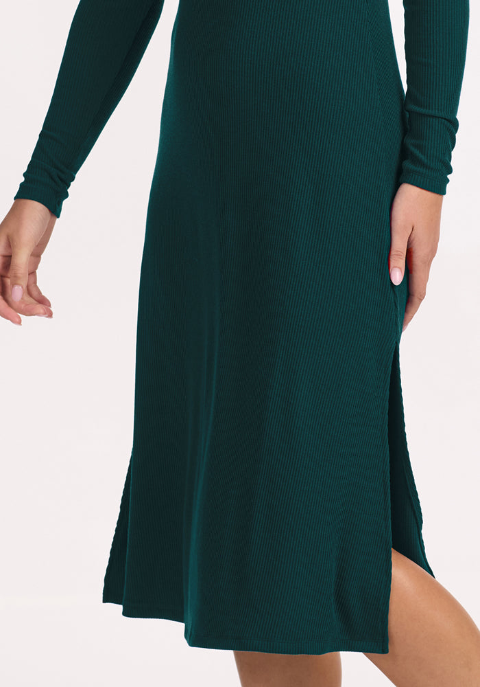 Someone wearing the Woolx Caroline Ribbed Twirl Dress in Deep Timber, a long-sleeve merino dress with a side slit, has their hand and leg visible against a plain light background.