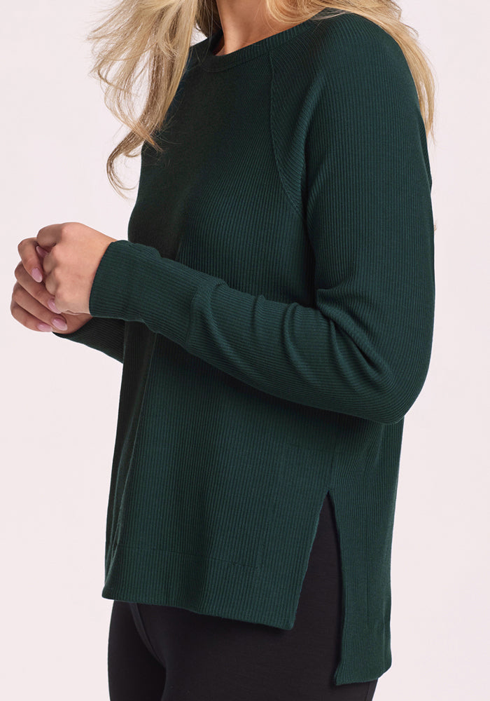A woman stands against a light background in the Woolx Ainsley Sweater - Deep Timber, a long-sleeved, dark green ribbed style with side slits, paired with black pants. Her hands are clasped, and her face is partially out of frame.