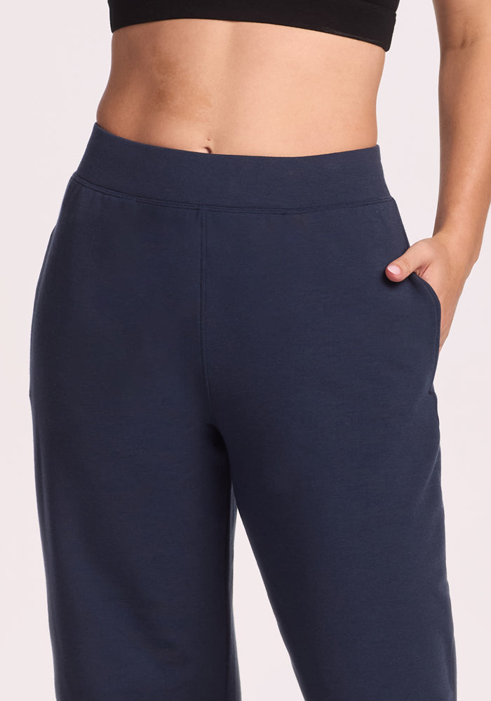 A person wearing Woolx Brynn Pants in Deep Navy and a black crop top places one hand in a pocket, creating a casual, relaxed look. The image highlights the waist and upper thighs.