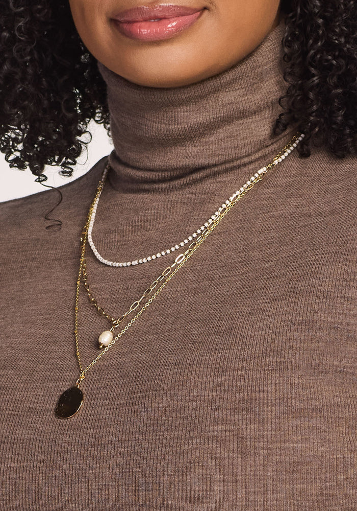 A woman wears the Woolx Sage Ribbed Turtleneck in Simply Taupe and four layered necklaces—beaded, two gold chains, and one pearl and gold pendant. Only her lower face and neck are visible.