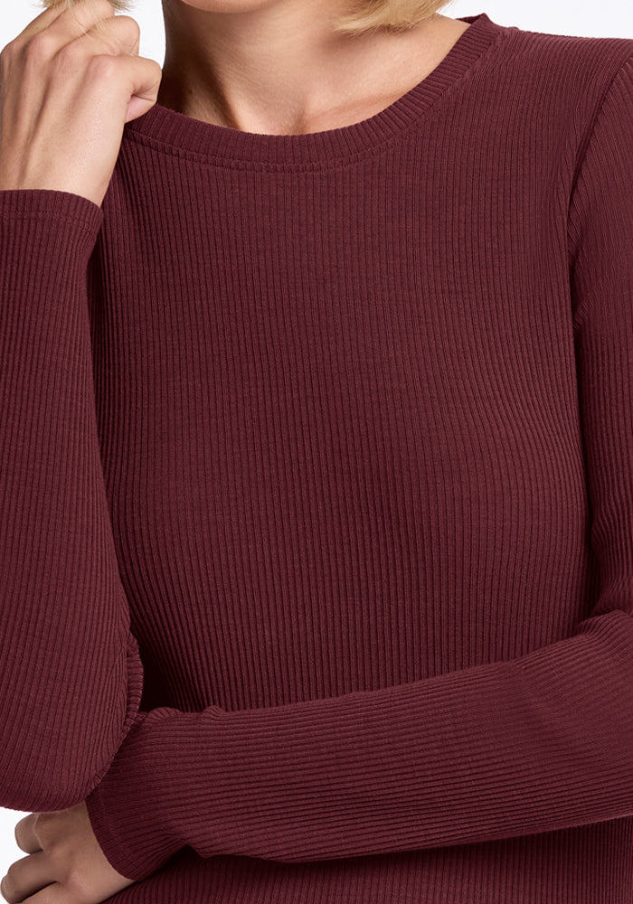 A person wearing the Woolx Hadley Ribbed Crew in Toasted Garnet, a long-sleeve women's merino wool top. The cropped image shows part of the face, arms, and upper torso—perfect for lightweight layering on travel days.