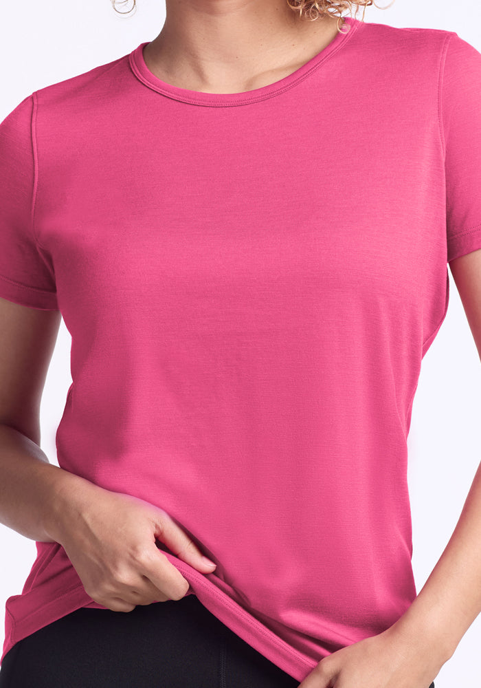 A person wearing the Woolx Liza Crew Neck Tee in Raspberry Fizz and black pants is holding the hem of the shirt with one hand. The image displays only their upper body.