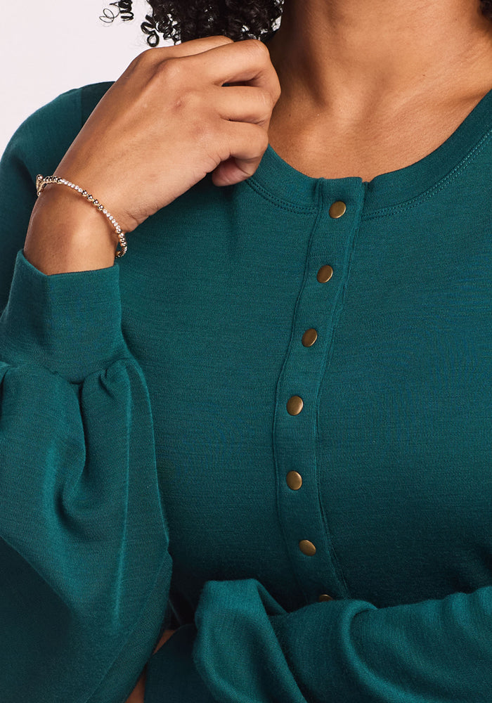 A person wearing the Woolx Charlotte Puff Sleeve Top in Riverbank Green and a delicate bracelet touches their chin, revealing part of their face and curly hair.
