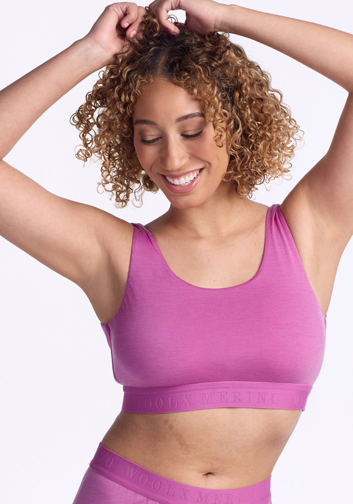 A smiling woman with curly hair wears the Woolx Aubrey Lounge Bra in Wild Blossom with matching bottoms, arms raised above her head, and stands against a plain white background.