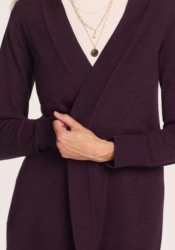 A person wearing the Woolx Macie Sweater in Deep Plum over a light top holds the front of the cozy cardigan closed with one hand, accessorized with layered gold necklaces.