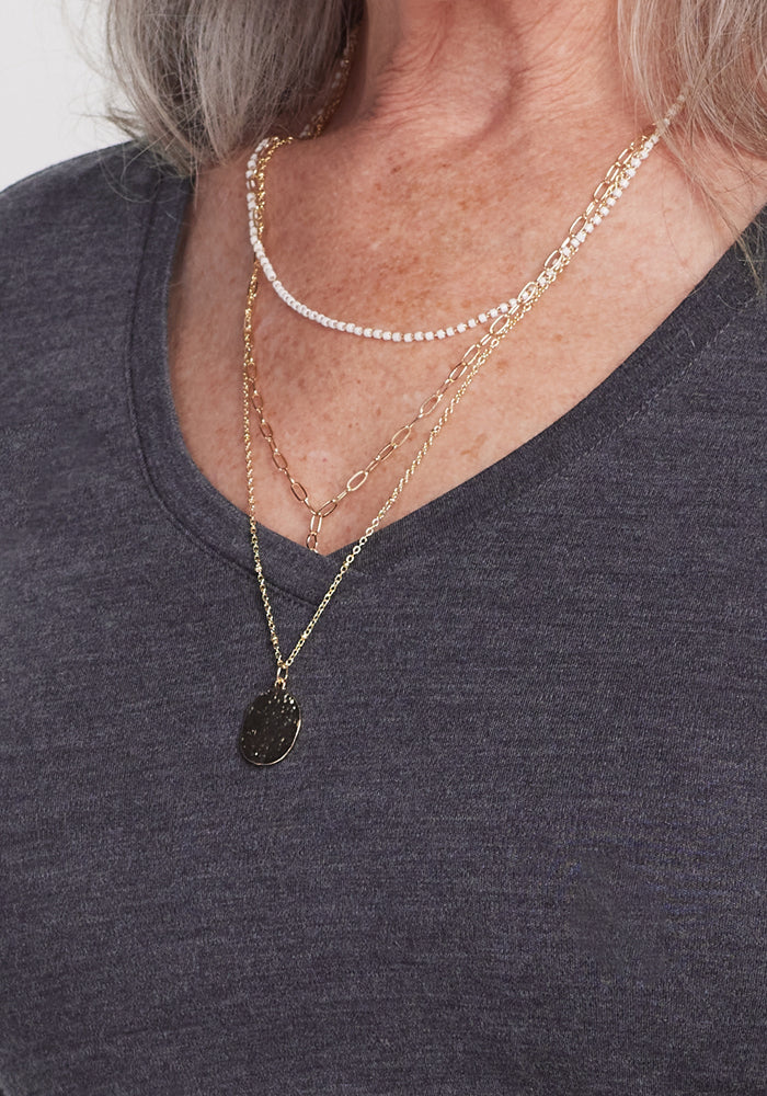 A close-up of a person wearing layered gold and pearl necklaces with a round pendant over the Woolx Eva Tunic in Cinder Melange—an elegant winter essential. The person's skin and some gray hair are also visible.