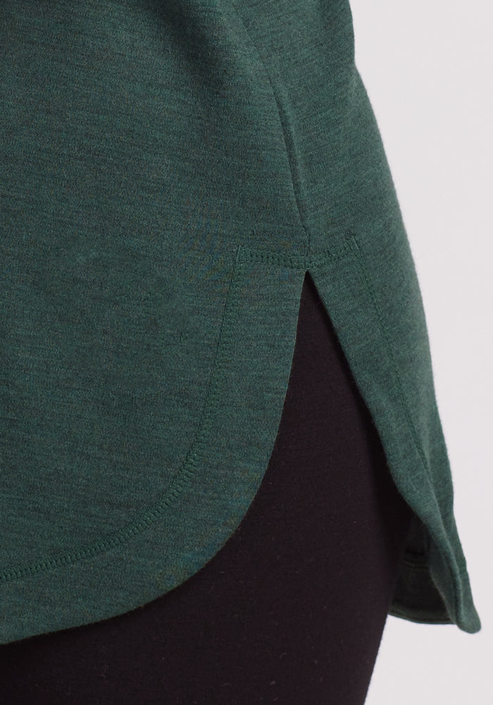 Close-up of the lower left side of the Woolx Blair Tunic in Forest, Merino wool with a rounded hem and side slit, offering lightweight warmth and layered over black pants.