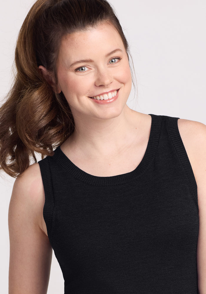 A woman with long brown hair in a ponytail smiles at the camera, wearing the Woolx Chessy Tank Top in black—a perfect layering piece—against a plain, light background.