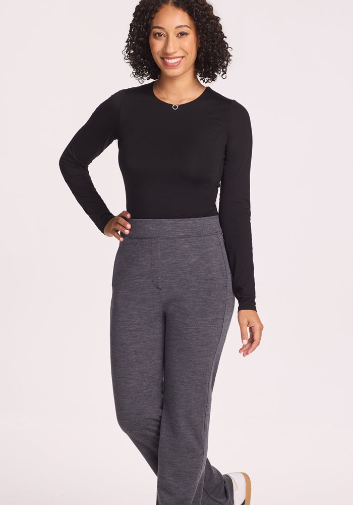 A woman with curly hair smiles, standing with one hand on her hip against a light background. She wears a long-sleeve black top and Woolx Emerson Straight Leg Pants Petite in Cinder Melange.