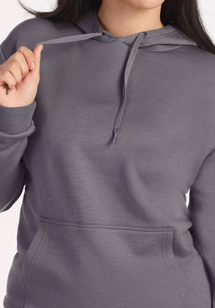 A person in a gray Woolx Avery Hoodie - Arctic Dusk holds a hoodie drawstring with their right hand. This Merino wool sweatshirt offers year-round comfort, has a front pocket, and the person's face is partially visible.