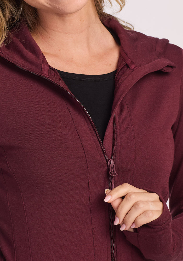 A person with neatly manicured nails is partially unzipping the Woolx Zoey Zip Up Hoodie in Toasted Garnet over a black top, showing only their lower face, neck, and upper torso.