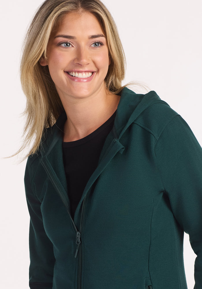 A smiling woman with blonde hair wears the Woolx Darby Zip Up Hoodie in Deep Timber over a black top, looking slightly to the side against a plain, light background.