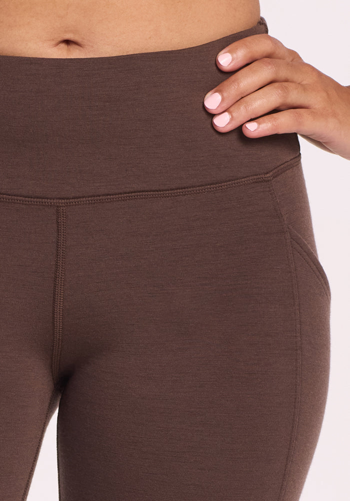 Close-up of a person wearing Woolx Piper Pocket Petite leggings in Bracken, featuring visible stitching and a side pocket. Only the lower torso and right hand are shown, with one hand resting on their hip.