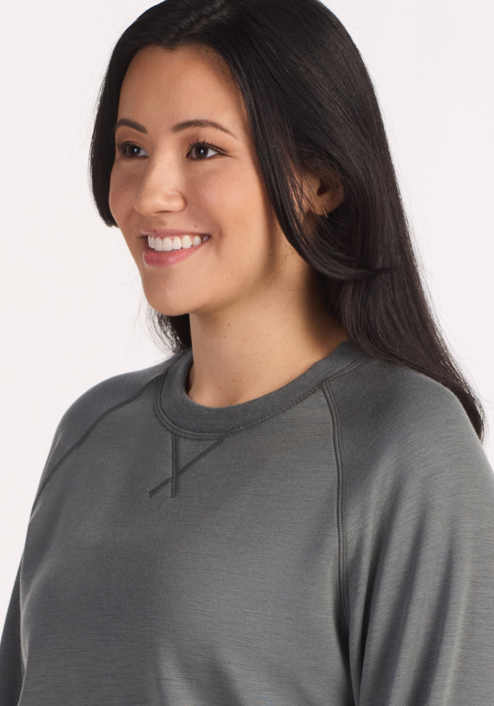 A woman with long, straight black hair smiles and looks to the side, wearing the Woolx Bailey Crewneck Sweatshirt in Sedona Sage, featuring visible chest and shoulder stitching. The background is plain white.
