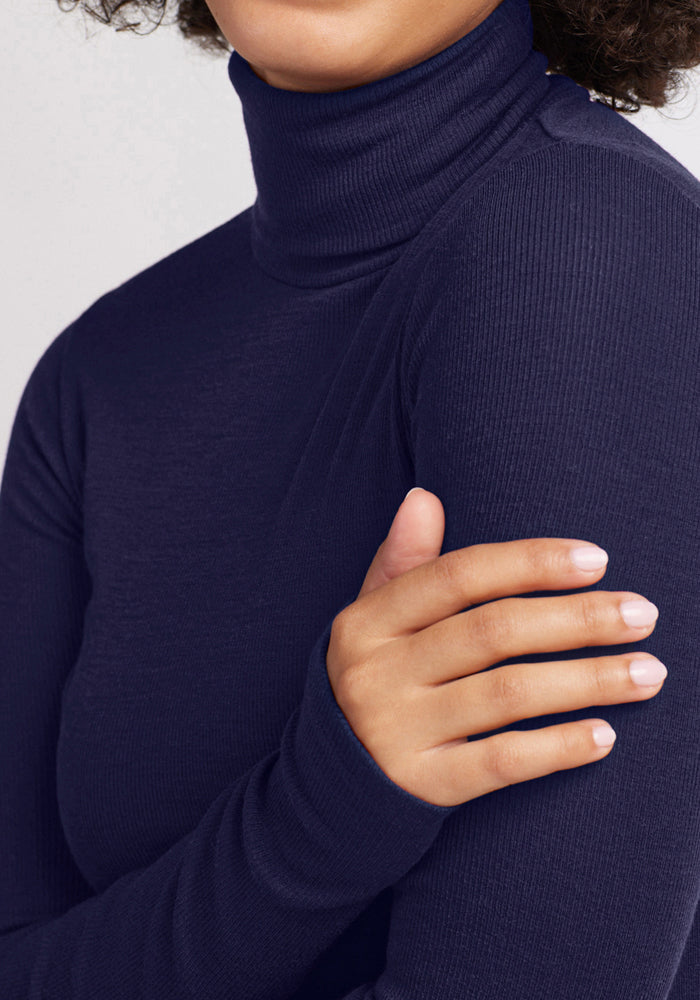 A person wearing the Woolx Sage Ribbed Turtleneck in Midnight Sapphire holds their left arm with their right hand, showing only the lower half of their face, neck, and upper torso.