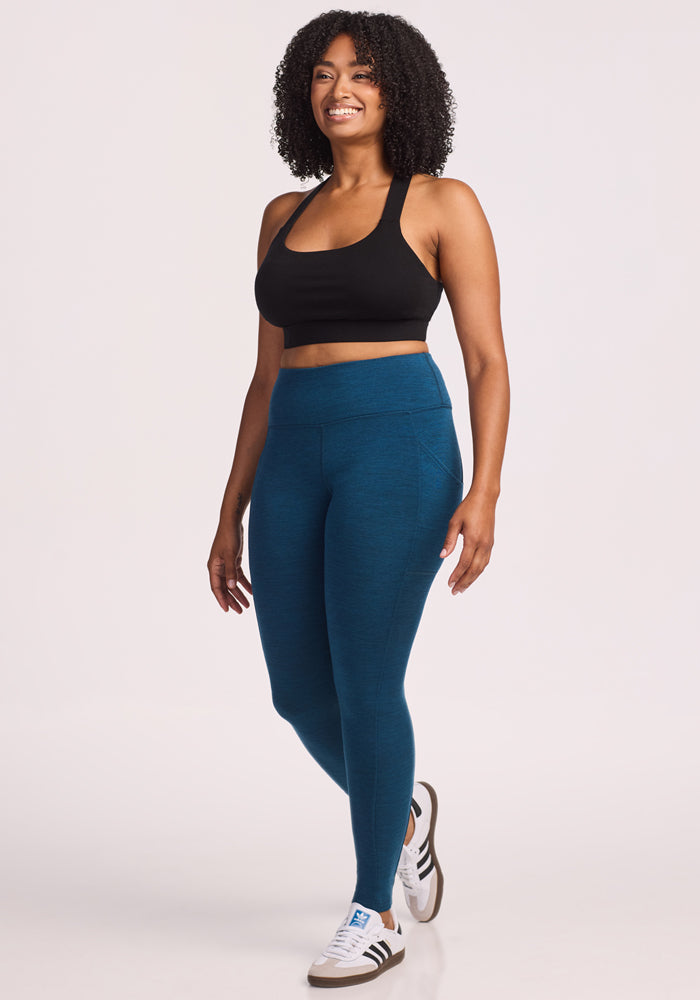 A woman with curly hair, in a Woolx black sports bra, Piper Pocket Leggings in Majolica Blue, and white sneakers, smiles against a plain white background.