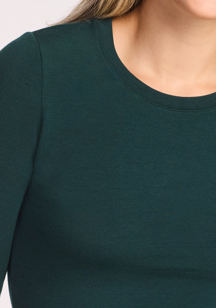 A close-up shows a person wearing the Woolx Sadie Baselayer Top in Deep Timber, made from soft Duralite merino. The image focuses on the neck, shoulder, and chest; the wearer’s face is partially out of frame.
