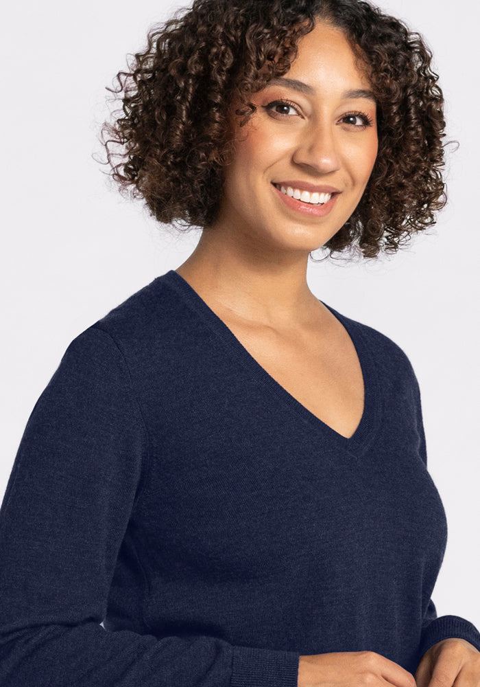 A woman with short curly hair smiles at the camera, wearing the Woolx Francesca Sweater in Storm Cloud. She stands against a plain light background, showcasing this travel-friendly merino wool sweater's comfort and style.