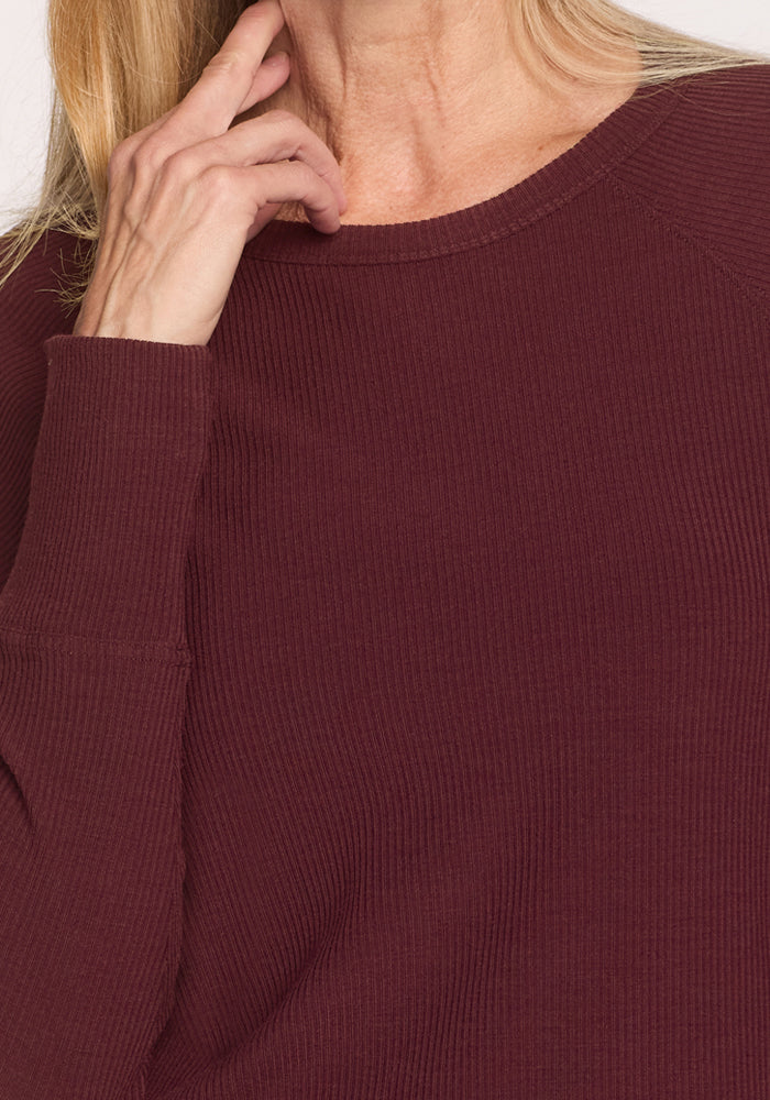 A close-up shows a person wearing the Woolx Ainsley Sweater in Toasted Garnet, featuring ribbed, long sleeves. Only the lower face and upper torso are visible as their hand rests near their chin.