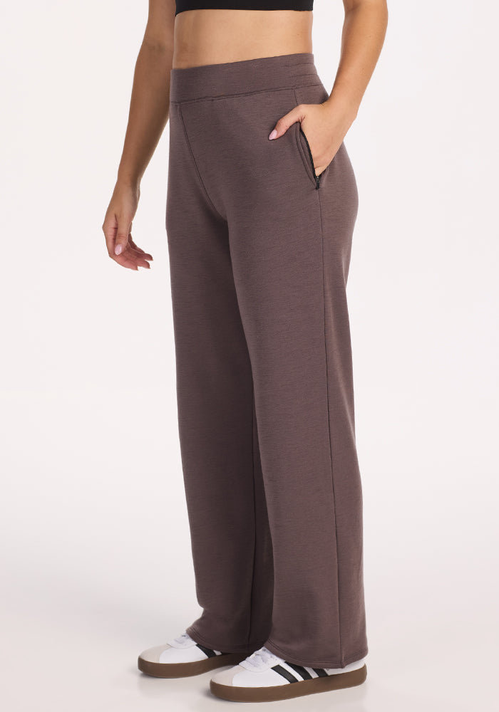 A person wears Woolx Bree Lounge Pants in Plum Truffle—high-waisted, straight-leg with side pockets—styled with a black crop top and white sneakers with black stripes, standing against a plain white background.