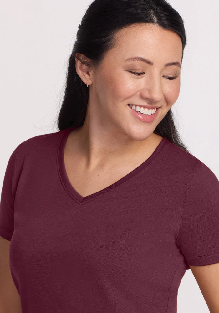 A woman with long dark hair smiles and looks down, wearing the Woolx Junie V Neck in Grape Harvest, a short-sleeved moisture-wicking shirt set against a light background.
