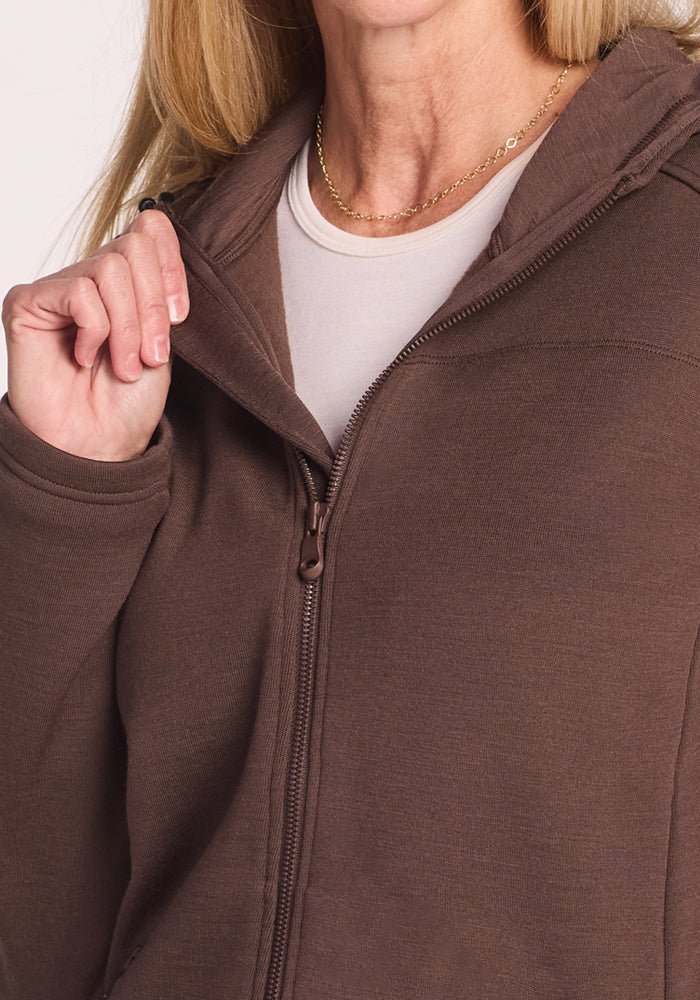 A person with long blonde hair wears the Woolx Cubby Hooded Sweatshirt in Bracken over a white shirt, holding the zipper partly open to show a gold chain necklace. The face is partially out of frame.