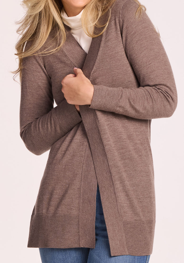 A woman with long blonde hair smiles while wearing the Woolx Macie Sweater in Simply Taupe over a white turtleneck and blue jeans, holding the cozy cardigan closed at the front with her face partially out of frame.