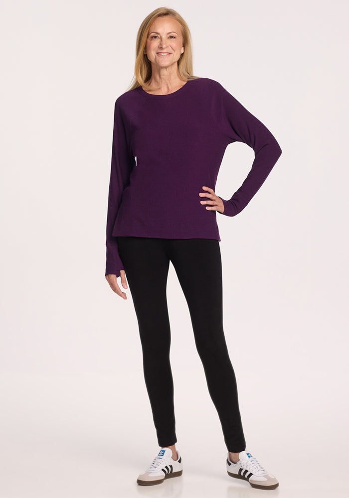 A woman with long blonde hair smiles in casual attire, wearing the Woolx Ainsley Sweater in Majestic Purple, black leggings, and white sneakers with black stripes. She poses confidently against a plain light background.