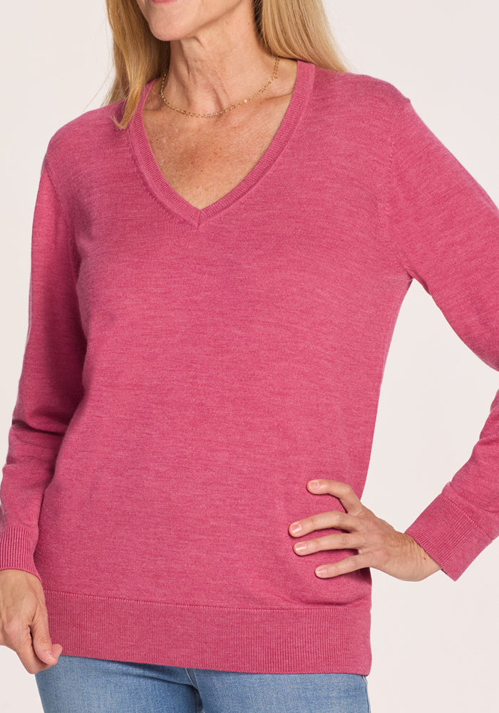 A woman models the Woolx Francesca Sweater in Wild Salmon with blue jeans, striking a pose against a plain background. Her face is partially out of frame.
