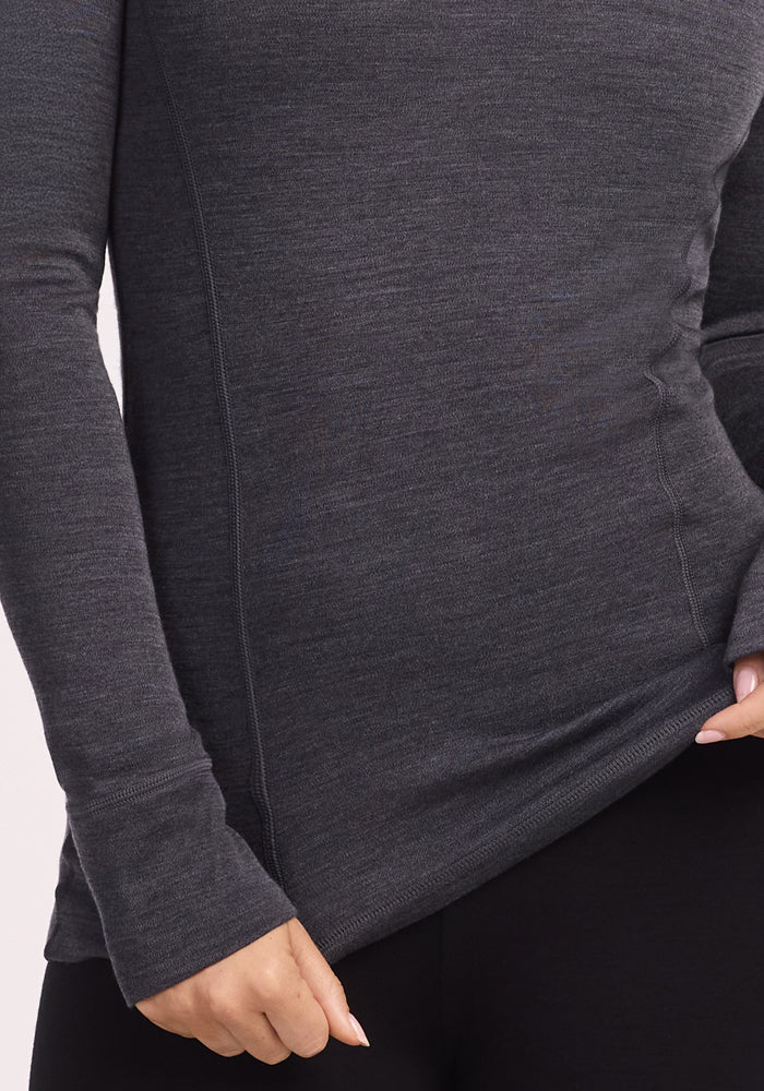 A person models the Woolx Hannah Long Sleeve Top in Cinder Melange with black pants, holding the shirt’s hem slightly out. Their face is not shown.