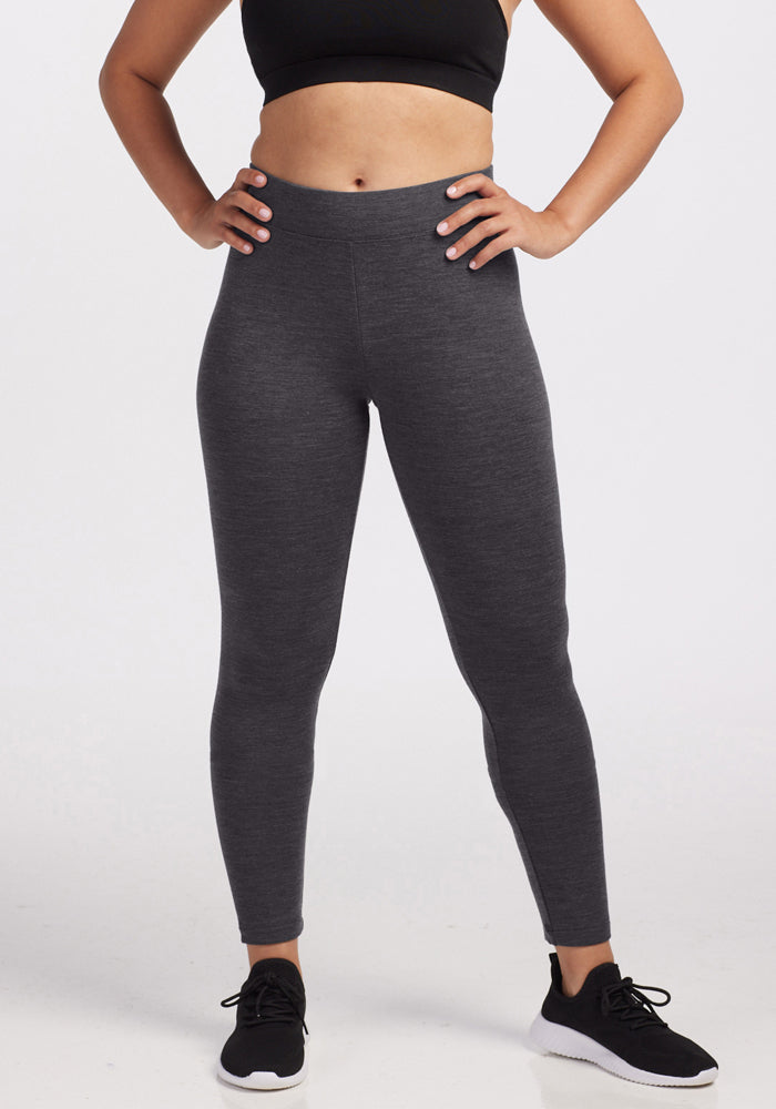 A person wearing a black sports bra, black athletic shoes, and Woolx Stella Leggings in Cinder Melange stands with hands on hips against a plain white background.