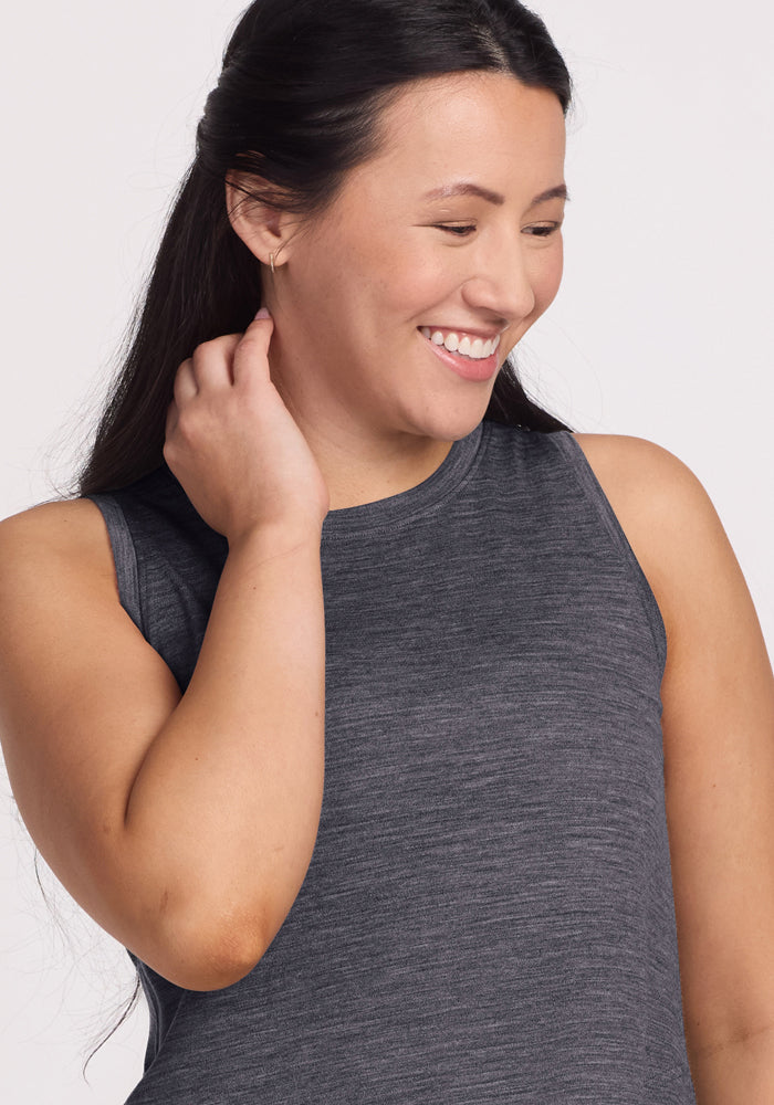 A woman with long dark hair smiles and looks down, touching her hair with one hand, while wearing the Woolx Clara Dress in Cinder Melange. The background is plain and light-colored.