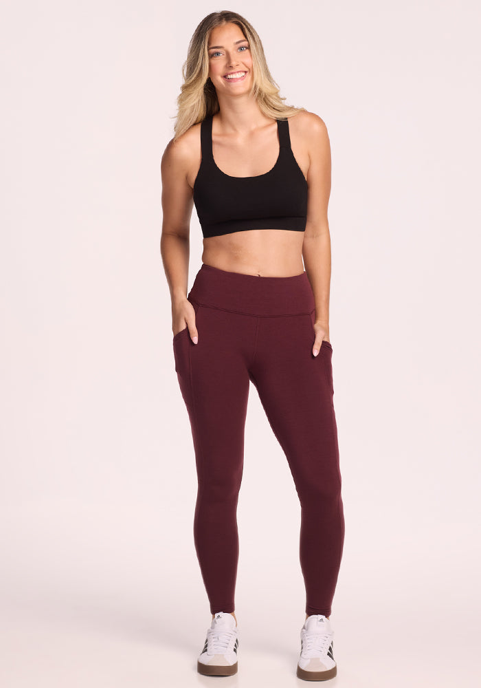 A woman with long blonde hair smiles while wearing the Woolx Piper Pocket Petite leggings in Toasted Garnet, a black sports bra, and white sneakers, posing with her hands in her pockets against a plain light background.
