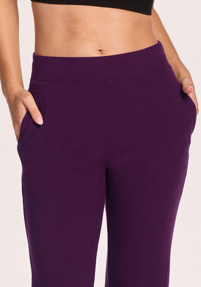 Model wearing Woolx Luca Ribbed Pants in Majestic Purple, high-waisted with ribbed texture and pockets, paired with a black crop top. Shown from mid-torso to mid-thigh against a plain background.