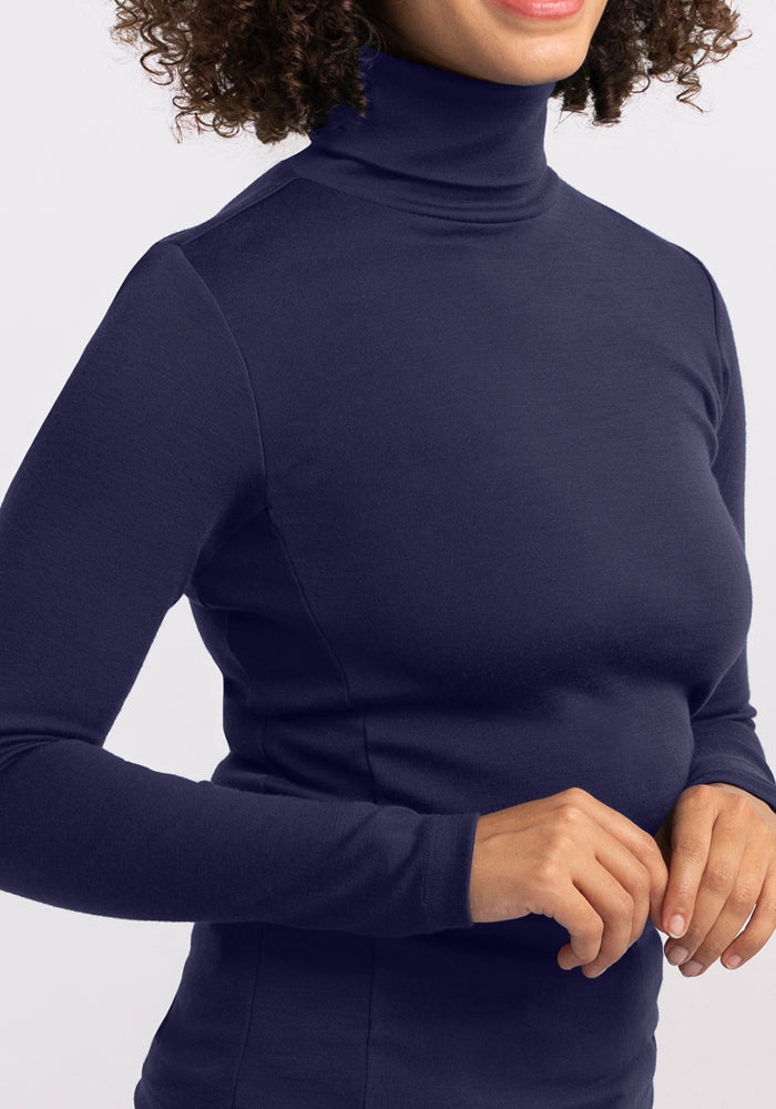 A person with curly hair wears the Woolx Peyton Turtleneck in Midnight Sapphire. Their face is partially out of frame, and their hands are clasped together in front of them.