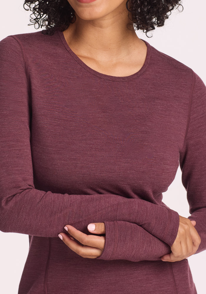 A woman crosses her arms, showing off neat light pink nails, while wearing the Woolx Elsa Long Sleeve in Grape Harvest Melange. The maroon merino wool top features thumb holes; her head is partially out of frame.