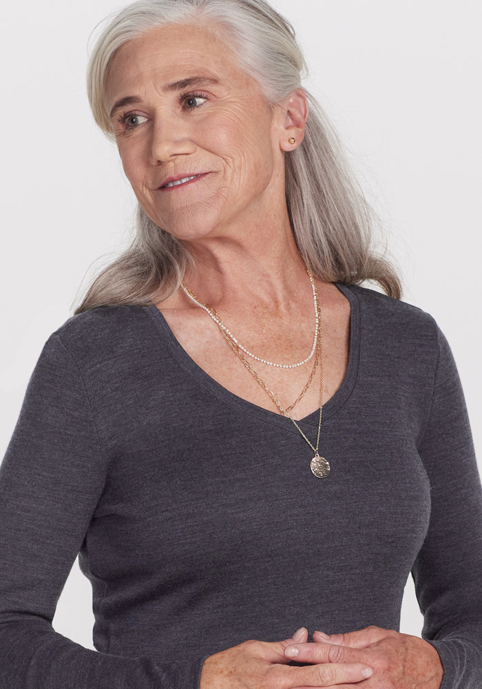 An older woman with long gray hair wears the Woolx Eva Tunic in Cinder Melange—an ideal winter wardrobe essential—layered with gold necklaces, gazing gently to the side against a plain white background.
