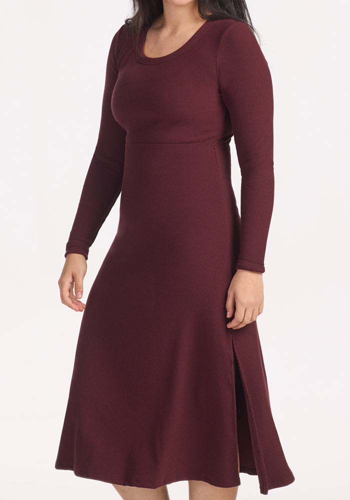 A person models the Woolx Caroline Ribbed Twirl Dress in Toasted Garnet, featuring long sleeves, a round neckline, and a side slit, highlighting its versatile silhouette against a plain white background.