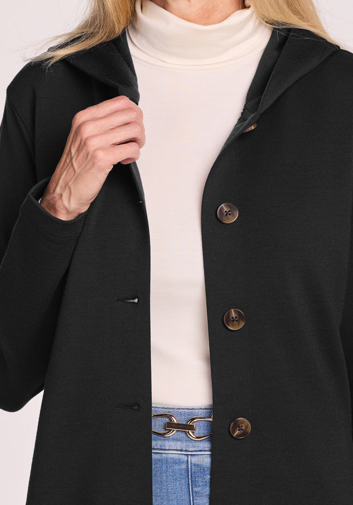 A person wears the Woolx Edna Hooded Cardigan in black over a white turtleneck and blue jeans with a gold chain belt, holding the cardigan open with one hand. Only their torso and part of their face are visible.