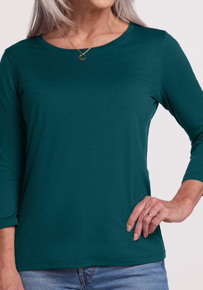 A woman with long gray hair wears the Woolx Jenny 3/4 Sleeve Crew Neck in Riverbank Green and blue jeans, standing with one hand on her hip. She accessorizes with a gold necklace and a ring.