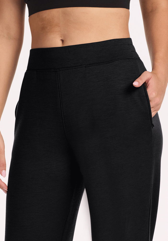 A person models Woolx Bree Lounge Pants in black, featuring a cozy straight-leg, high-waisted fit, with one hand in the pocket; shown from mid-torso to mid-thigh against a plain background.