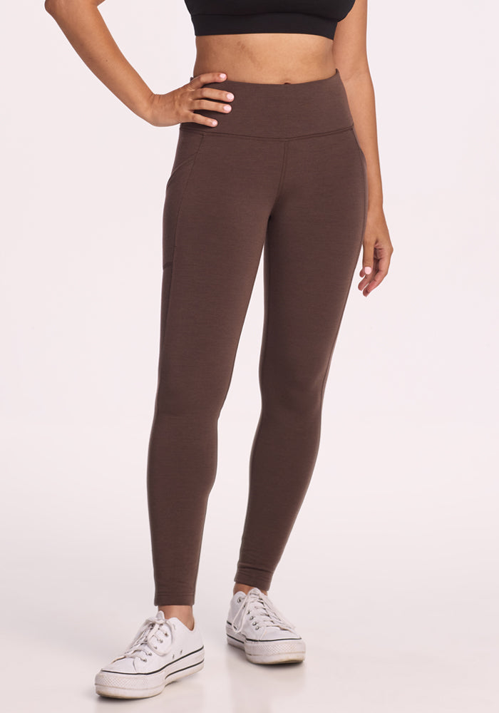 A person wears Woolx's Piper Pocket Petite - Bracken high-waisted brown cold-weather leggings, a black crop top, and white sneakers against a plain light background.