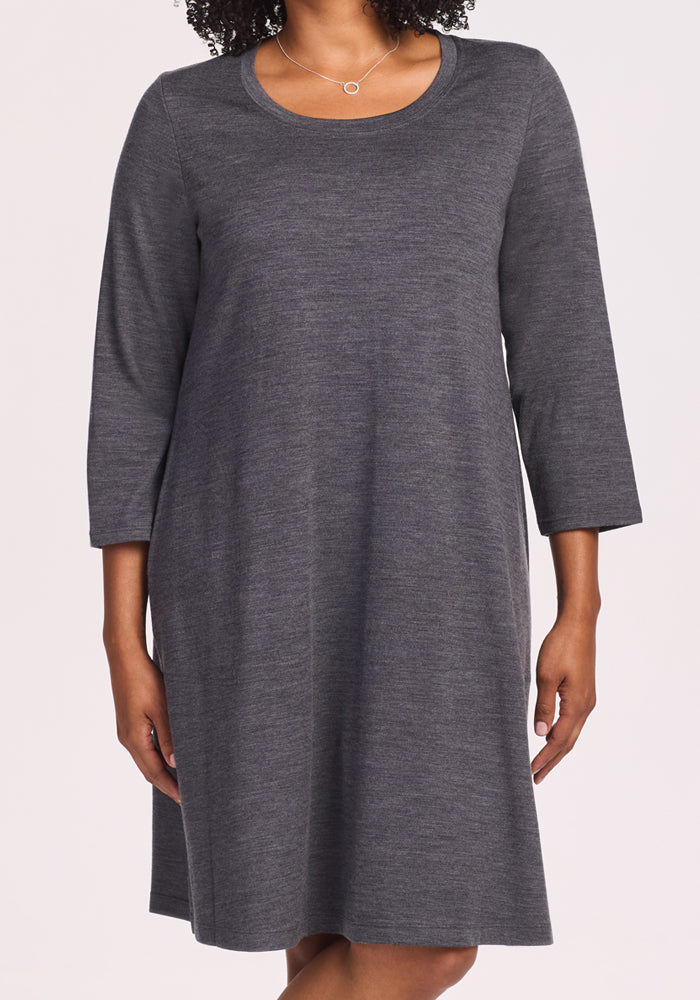 A woman stands against a plain light background wearing the Woolx Rissa A Line Dress in Cinder Melange—a simple Merino wool dress with three-quarter sleeves and a round neckline, ideal for layering in fall and winter.