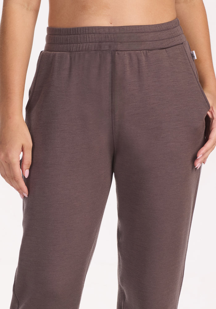 A person stands with hands in pockets against a white background, wearing Woolx Parker Sweatpants in Plum Truffle—high-waisted, loose-fitting Merino wool lounge pants with an elastic waistband for optimal temperature regulation.