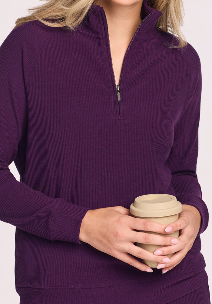 A person in the Woolx Quinn Ribbed 1/4 Zip in Majestic Purple holds a beige reusable coffee cup with both hands, showing only the lower part of their face and upper body.
