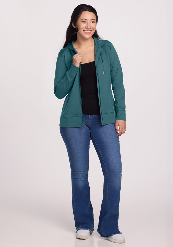 A woman with long dark hair smiles against a light background, wearing the Woolx Ryann Hoodie in Tidepool—a teal merino wool essential perfect for travel—paired with a black top, blue jeans, and white sneakers.