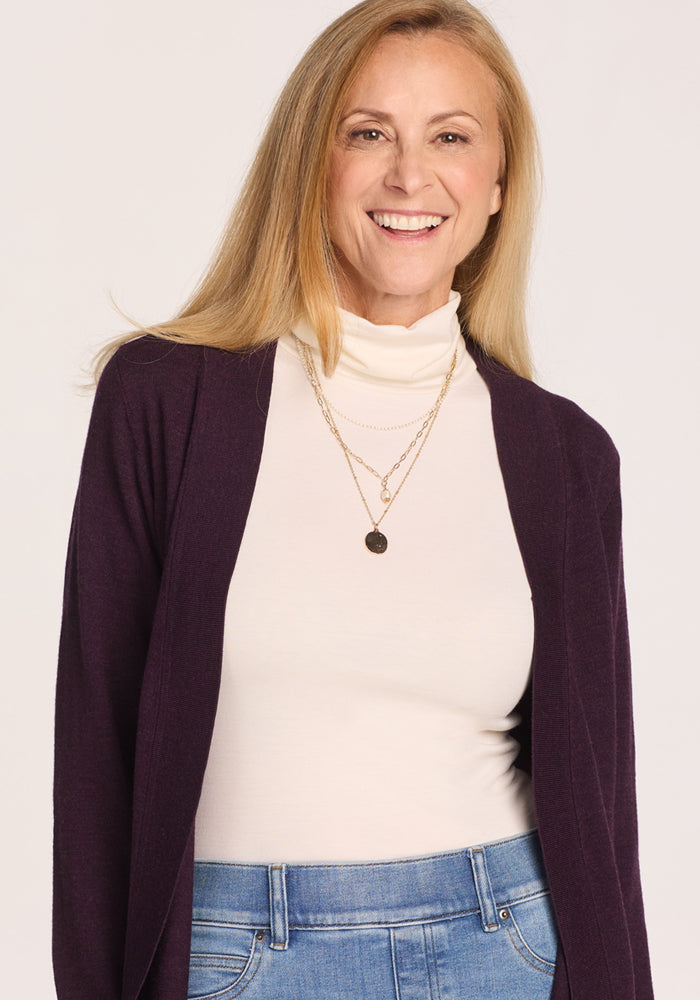 A smiling woman with long blonde hair wears a white turtleneck, blue jeans, and the Woolx Macie Sweater in Deep Plum—a merino wool open-front cardigan—while standing against a plain light background.
